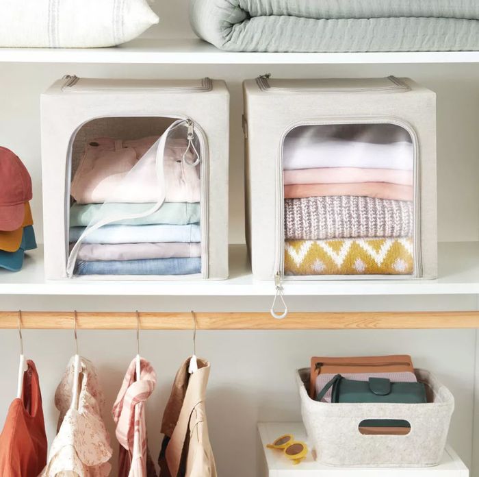 Folded clothes inside beige storage bins on a shelf, with clothes and hats hanging close by.