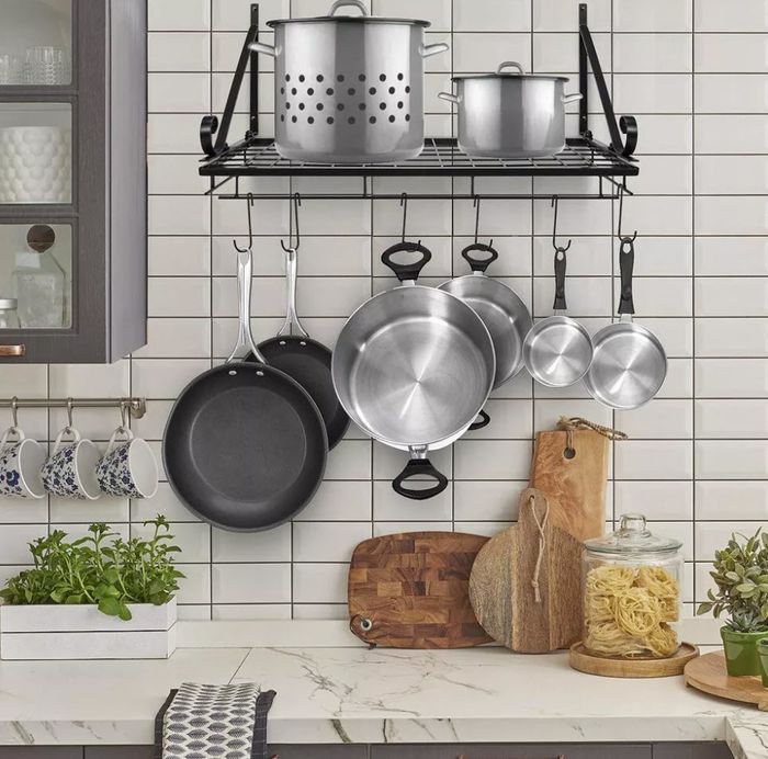 A kitchen featuring a wall-mounted pot rack, hanging cookware, cutting boards, plants, and a pasta jar on the counter, with a modern tiled backsplash for added flair.