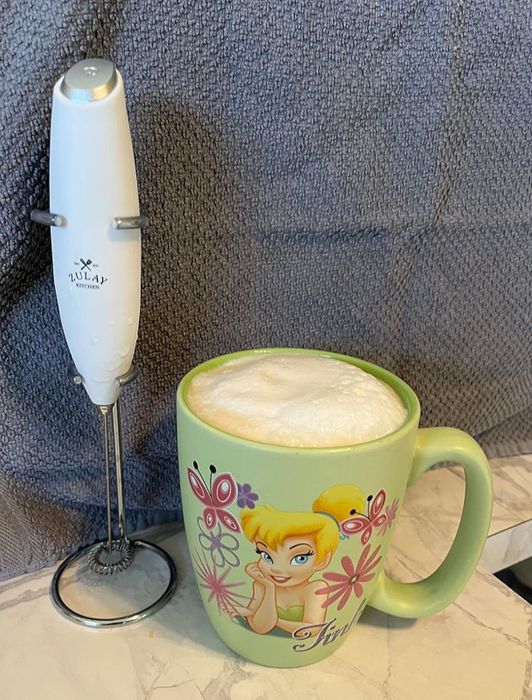 An electric frother placed beside a Tinker Bell mug filled with frothy milk, sitting on a kitchen counter