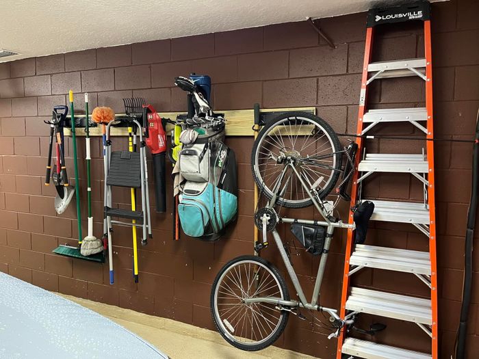 Various items such as a golf bag, bicycles, and a ladder are arranged on a garage wall using hanging storage solutions.