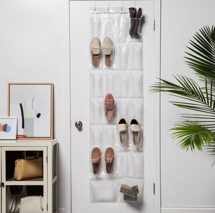 An over-the-door shoe organizer with shoes and a purse hanging, next to a small cabinet and a framed abstract painting. A plant can be seen in the corner.