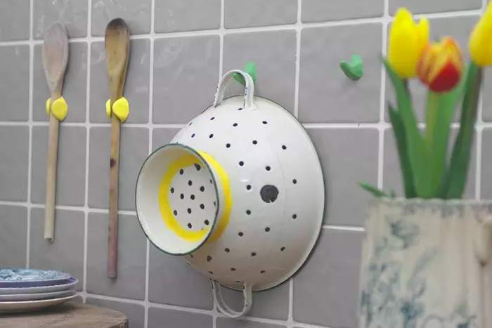 A colander and wooden spoon mounted on a tiled kitchen wall using hooks and holders made from moldable glue