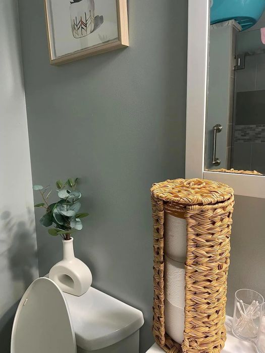 A bathroom with a wicker basket holding toilet paper, a small plant in a white vase, and a framed picture on the wall