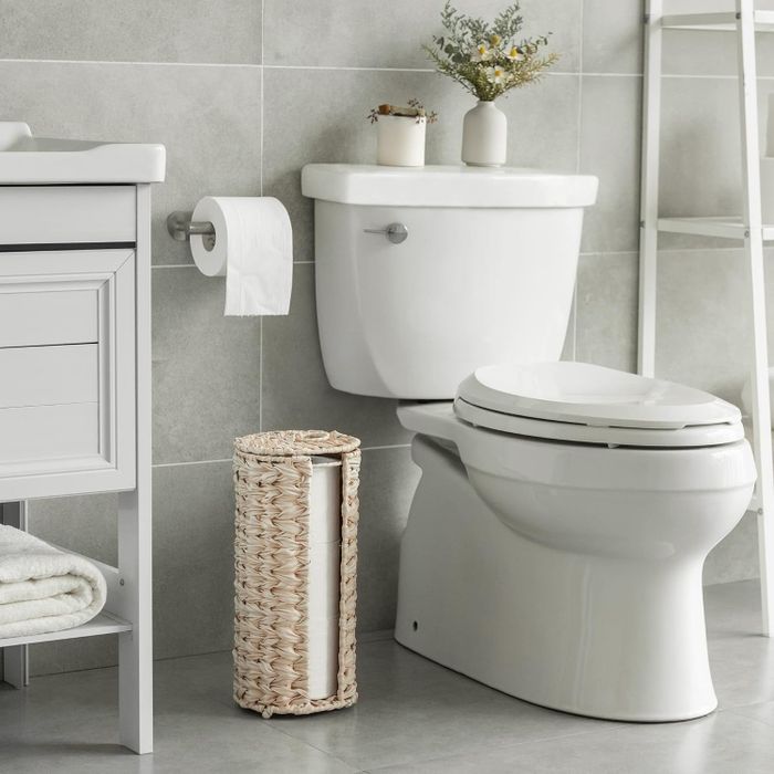A bathroom scene featuring a modern toilet, plants on the tank, a wicker basket filled with toilet paper, and towel storage next to the vanity.