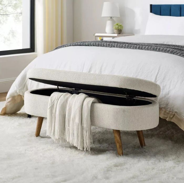 A white bedroom bench with wooden legs, partially opened to reveal a folded blanket inside, sitting at the foot of a cozy, modern bed.