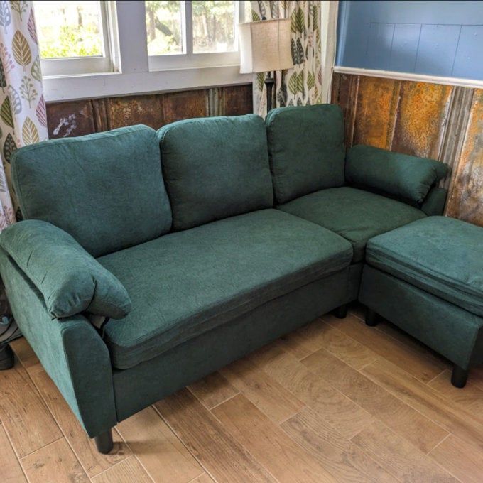 A green sectional sofa paired with an ottoman, situated in a warm, rustic room featuring wood-paneled walls and curtains adorned with leaf patterns.