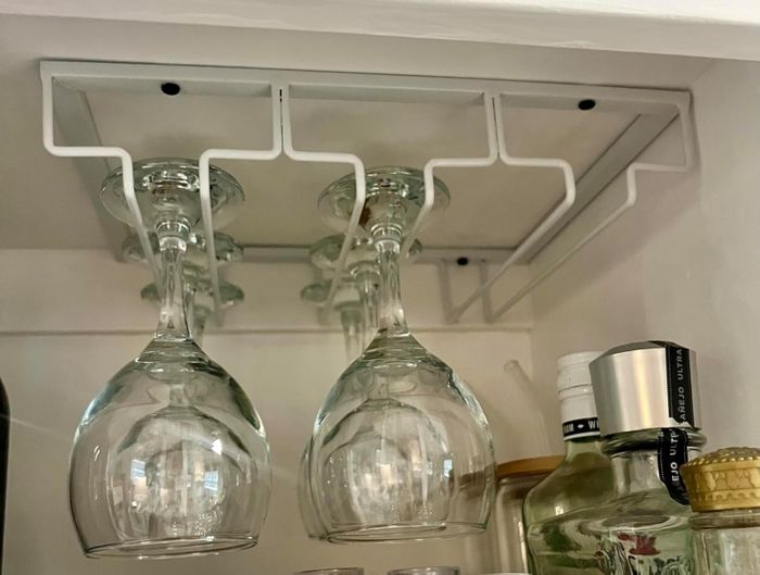 Wine glasses are neatly suspended upside down from a mounted rack inside a cabinet, with an assortment of bottles visible nearby, illustrating a smart and tidy kitchen storage solution.