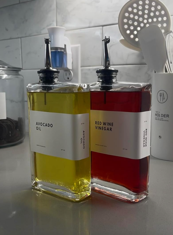 Two square glass bottles with pour spouts labeled “Avocado Oil” and “Red Wine Vinegar,” sitting on a kitchen counter