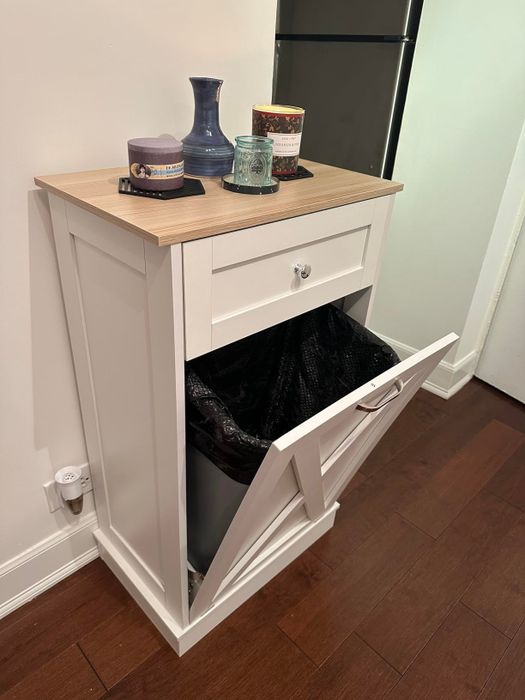 A small white kitchen cabinet with a wooden top, holding candles and a vase, and featuring a pull-out trash bin on the lower half