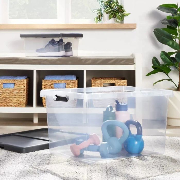 A clear plastic storage bin with handles holds a water bottle, two kettlebells, and two dumbbells. Nearby, a smaller bin filled with shoes rests on a bench.