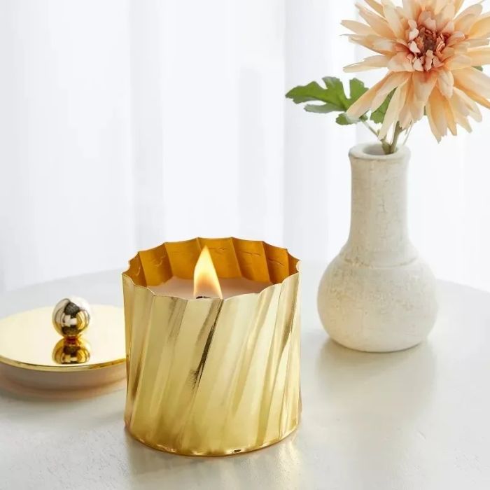 A lit candle with a decorative gold holder, placed next to a pink flower in a white vase, creating a cozy shopping aesthetic on a table.
