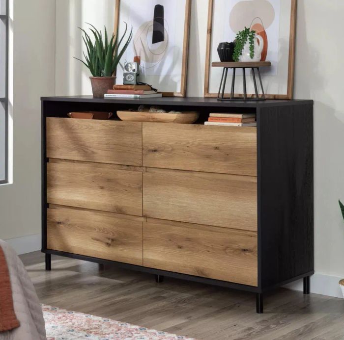 A sleek modern dresser featuring six wooden drawers. On top, there are two framed art pieces, a small plant, a clock, and decorative accents. Light pours in from the left side of the room.