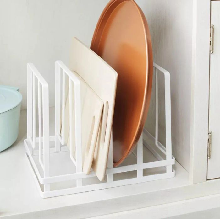 A white metal rack on a kitchen counter holds cutting boards and a large plate stored upright, neatly organized.