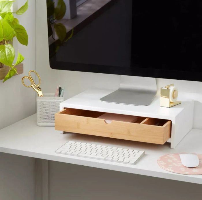 A well-organized desk featuring a large computer monitor, a keyboard, a potted plant, scissors, and a drawer beneath the monitor that holds a pink book.