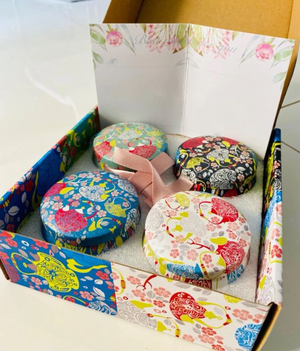 a photo of a reviewer showcasing an open box filled with four colorful and patterned tin candles