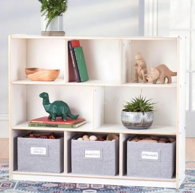 A white bookshelf with plants, a wooden bowl, books, toy dinosaurs, a plant pot, and fabric bins labeled