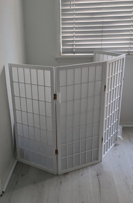 A three-panel white wooden folding room divider positioned in a minimalist room beside a window with blinds