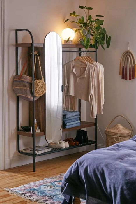 Bedroom corner with a modern clothing rack. Items include folded jeans, a beige sweater on a hanger, woven bags, and a mirror.
