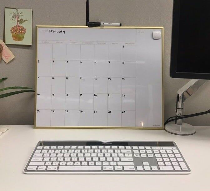 A desk setup featuring a keyboard, computer monitor, and a blank whiteboard calendar for February, with a pen clipped on top of it.