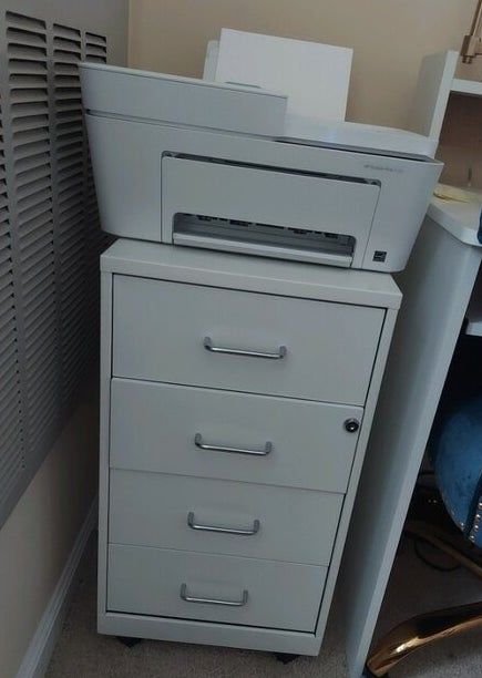 A grey printer placed on top of a four-drawer filing cabinet in an office environment. No people or text visible in the image.