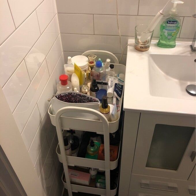 A bathroom storage cart stocked with toiletries such as bottles, creams, and sprays, positioned next to a sink with dish soap and a glass