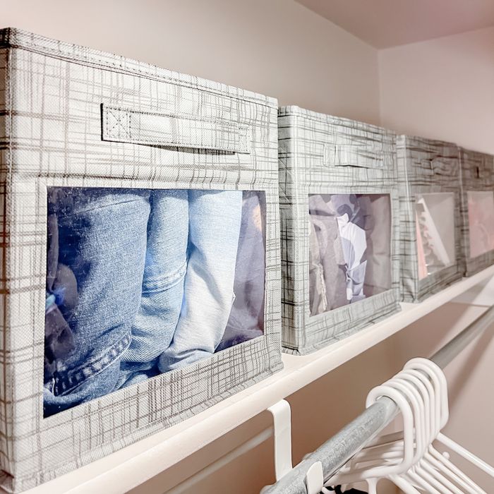 Three fabric storage bins with windows, each showing different folded clothes inside. Hangers are visible on the rail below.