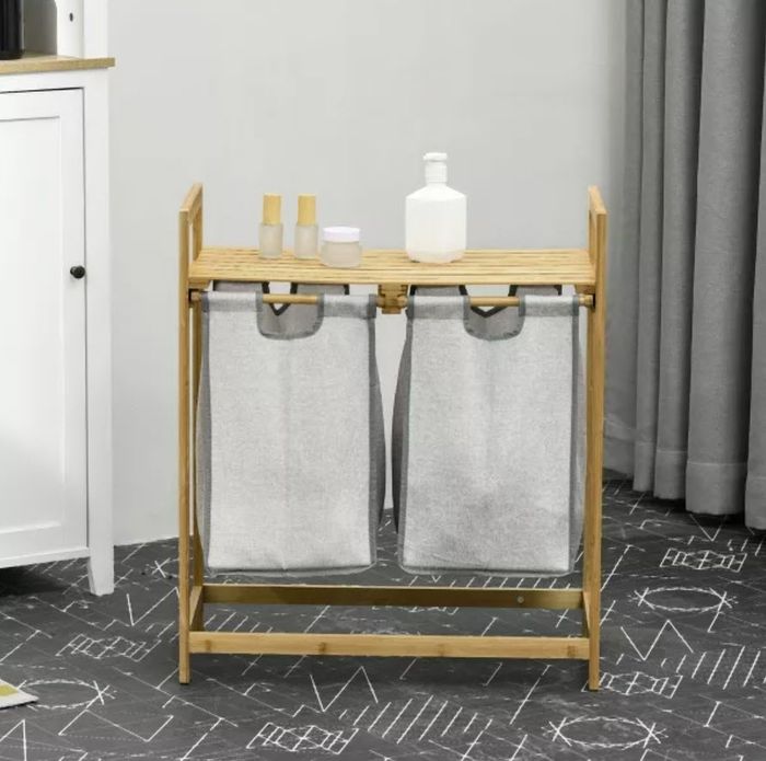 A laundry hamper stand featuring dual gray fabric bags, a wooden frame, and bottles placed on the top shelf, with drapes and a cabinet visible in the background.