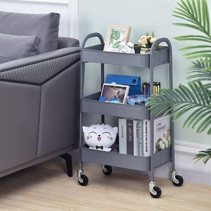 A grey three-tier rolling cart filled with items like books, pens, a tablet, a plush toy, and small decorative objects