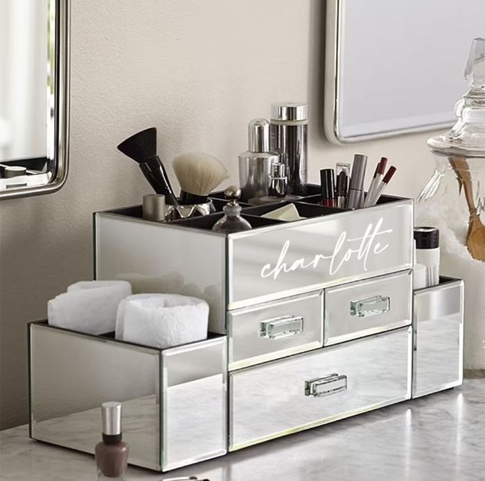 A mirrored beauty organizer with the label 'Charlotte' holds makeup items, brushes, and beauty tools, positioned on a marble countertop near a mirror.