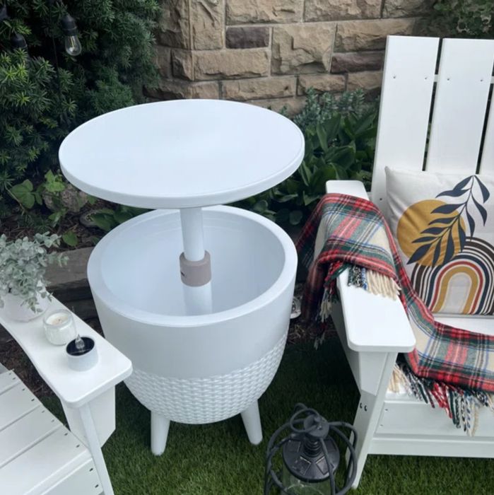 A white patio table with a built-in cooler, positioned between white Adirondack chairs—one adorned with a colorful blanket and pillow. A candle and small plant sit on the chair