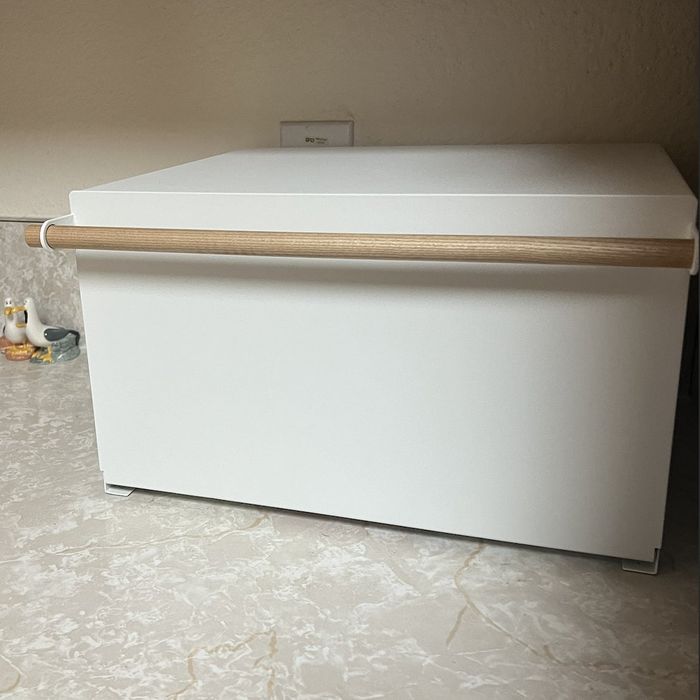 A sleek white bread box with a wooden handle, placed on a kitchen countertop beneath cabinets