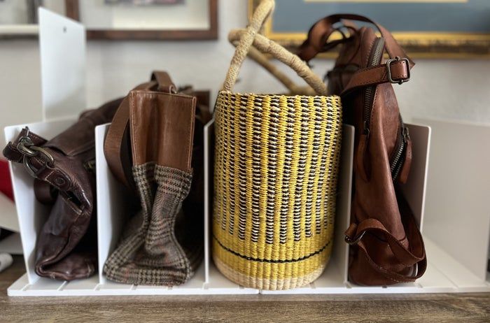 Four bags neatly arranged on a white shelf: two leather bags, a woven straw bag with rope handles, and a patterned fabric bag