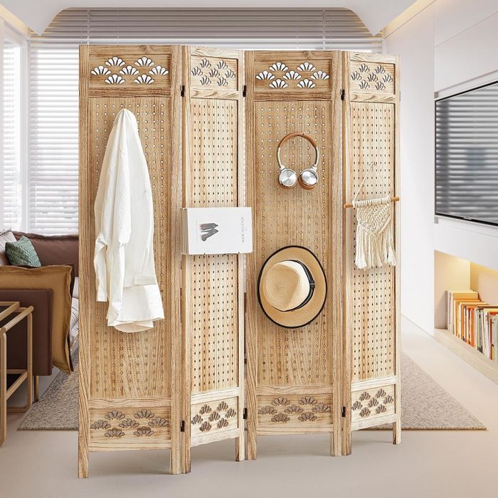 A wooden room divider with detailed cut-out patterns, decorated with a white shirt, a hat, a book, headphones, and a macramé wall hanging