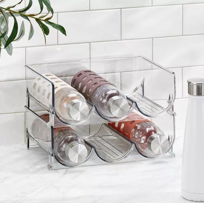 Four glass water bottles with metal caps neatly arranged in a clear plastic rack on a marble countertop next to a white insulated bottle.