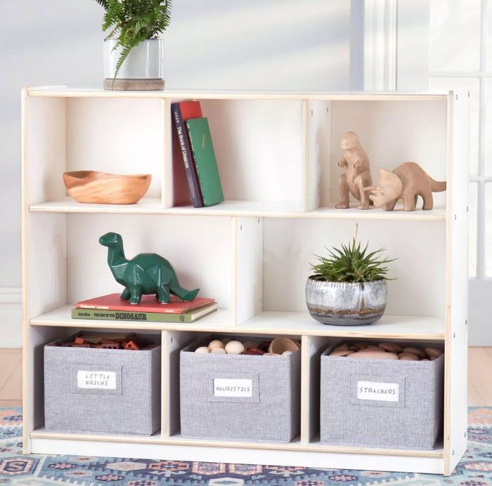 A wooden bookshelf stacked with a potted plant, books, toy dinosaurs, a wooden bowl, and three labeled gray storage bins: