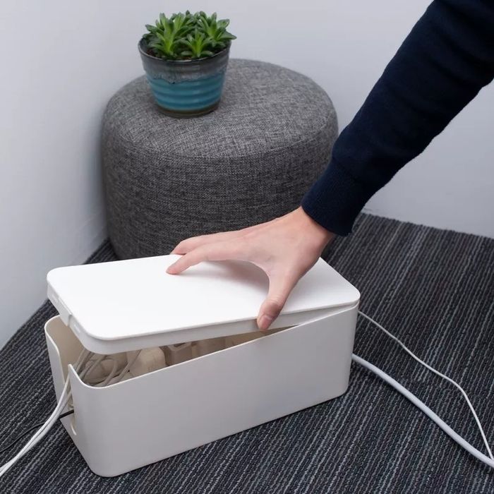 A hand places a white plastic cover on a cable organizer box beside a small gray ottoman with a potted succulent.