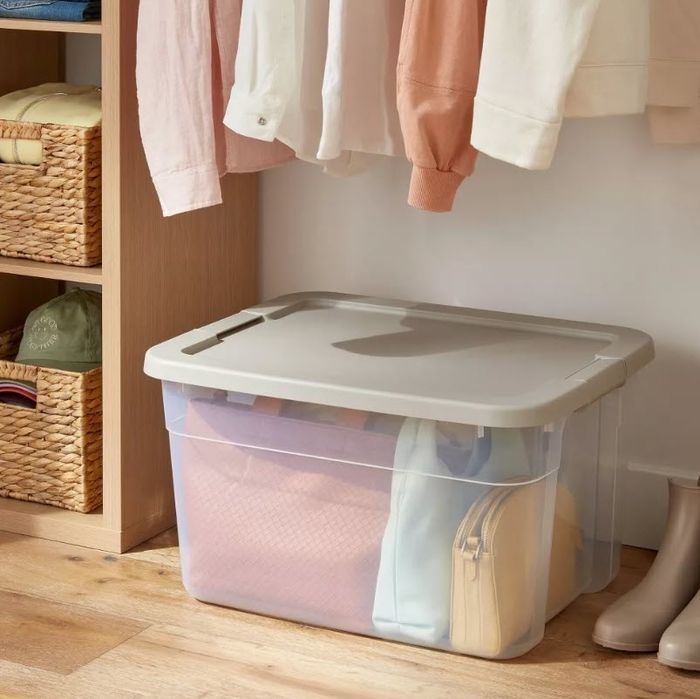 A plastic storage bin with a lid, filled with folded clothes and bags, placed on the floor of a closet with hanging clothes, and accompanied by nearby woven baskets containing more clothing.
