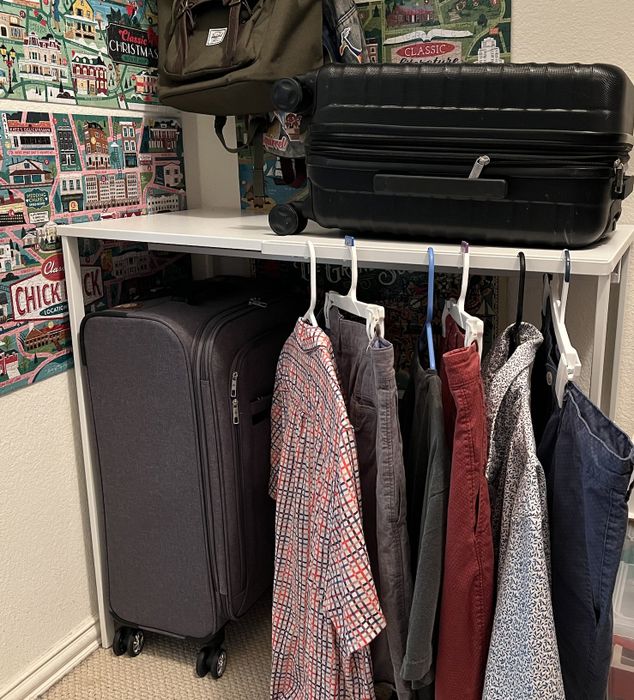 A small closet with neatly hung shirts and pants, two suitcases, and a bag resting on a shelf above, with Christmas village wallpaper adding a cozy touch to the walls