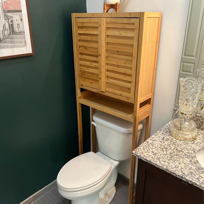 A reviewer's photo of a bamboo shelf positioned above a white toilet, next to a sink with glassware neatly arranged on a marble countertop.