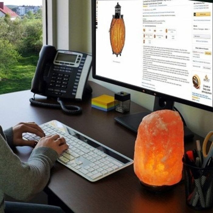 A person shopping online at a desk featuring a glowing Himalayan salt lamp, a telephone, a keyboard, and a monitor displaying a product page