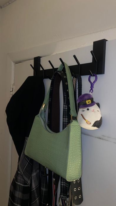 A wall-mounted coat rack displaying a green handbag, a scarf, a black clothing item, and a small plush toy with a purple hat and hook