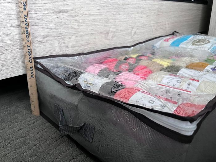 A storage container filled with assorted yarn, neatly organized and visible through a clear plastic lid, placed under a bed