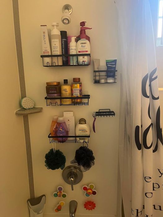 a reviewer's shower wall featuring five black adhesive shelves holding various shampoos, bottles, and two loofahs hanging from the hooks