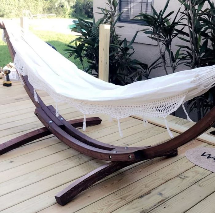 A wooden hammock stand supporting a white fringed hammock on a backyard deck, with plants in the background.