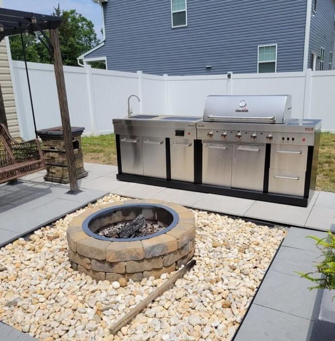 An outdoor kitchen setup featuring a sleek stainless steel grill, spacious countertops, and cabinets, paired with a circular stone fire pit surrounded by pebbles in a fenced backyard.