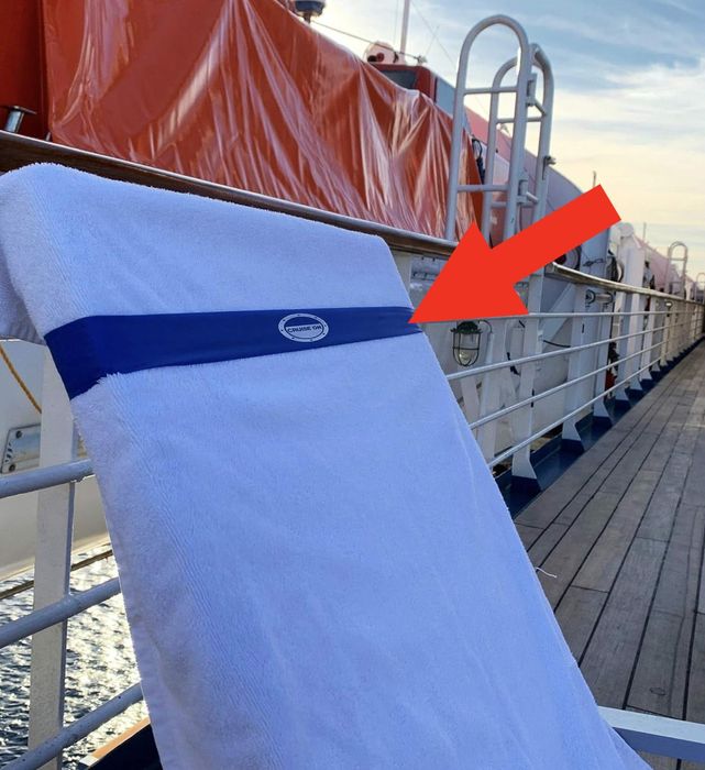A white towel with a logo draped over a ship’s railing, with the deck and lifeboats in the background.