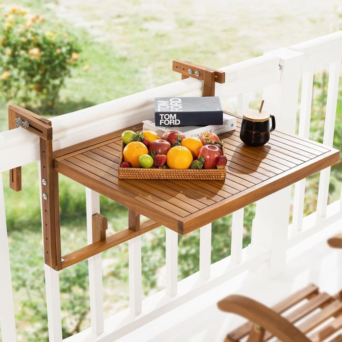 A wooden balcony table with a fruit basket, a book, and a cup resting on top, mounted on a white railing