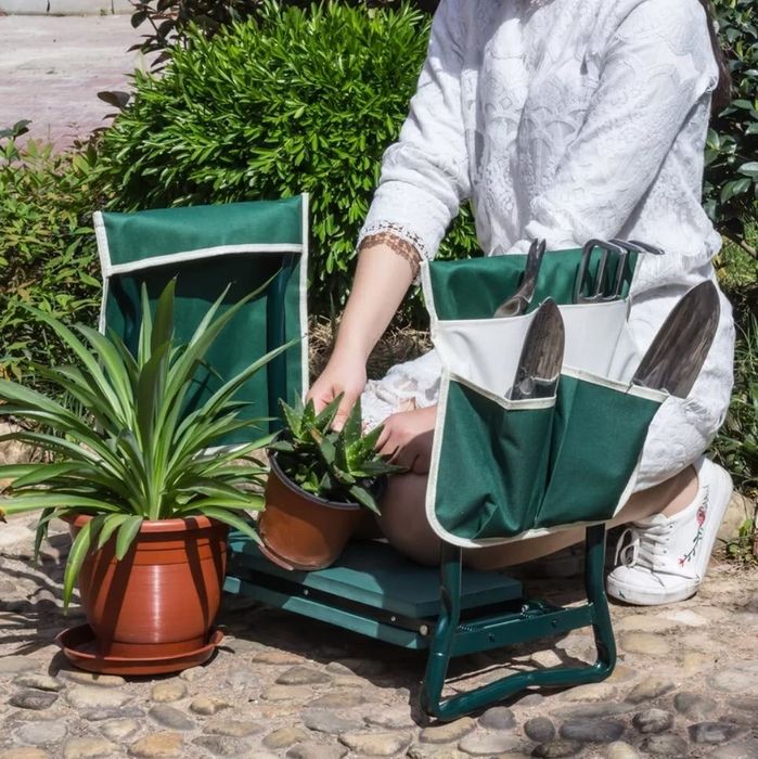 A person gardening while seated on a green garden stool with gardening tools stored in the pouches. They are planting a small potted plant next to a larger terracotta potted plant.