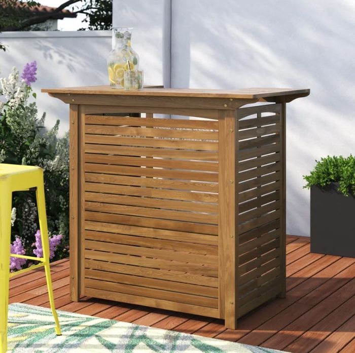 A wooden outdoor bar table with a pitcher of lemonade and glasses, accompanied by a yellow barstool and plants in the background, creating a welcoming outdoor bar scene.