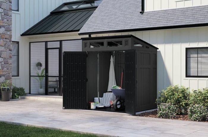 An outdoor storage shed with open double doors revealing gardening tools, buckets, and a hose inside. The shed is located next to a modern home featuring large windows and a patio.
